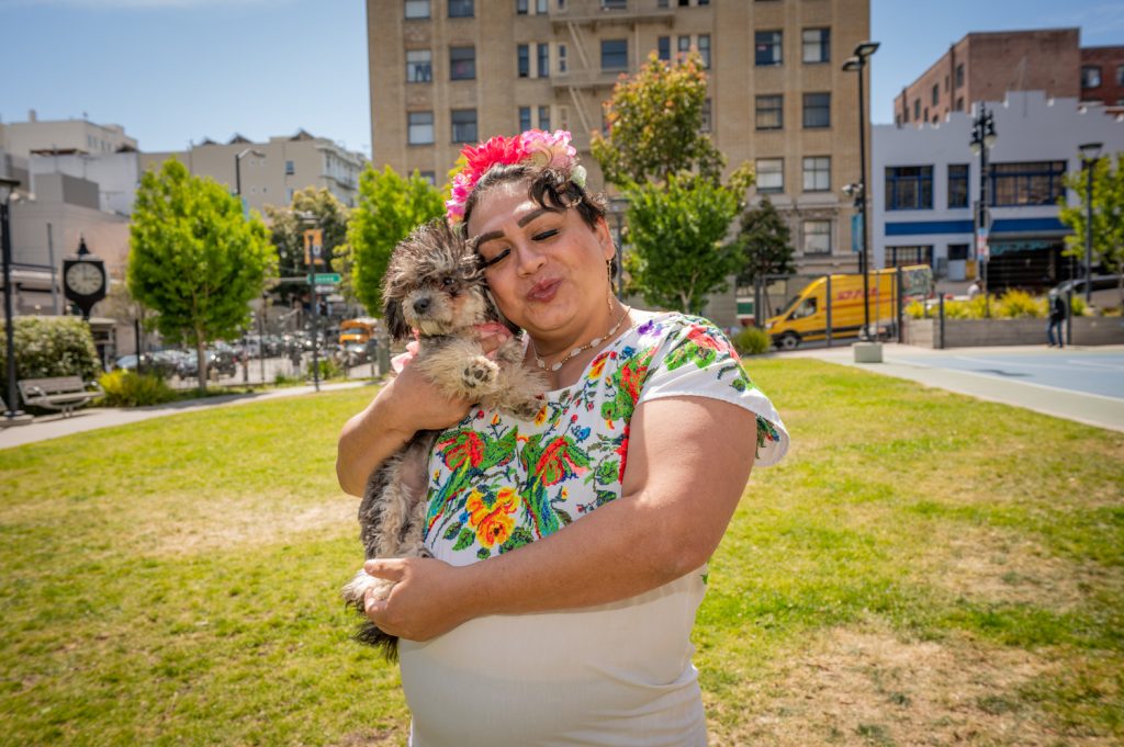 Johana (a woman) wearing a white dress with embroidered flowers and a flower headband, holding her small dog, standing in a park with buildings in the background.