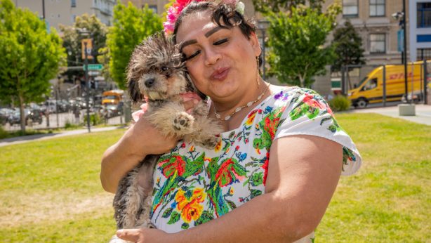 Johana (a woman) wearing a white dress with embroidered flowers and a flower headband, holding her small dog, standing in a park with buildings in the background.