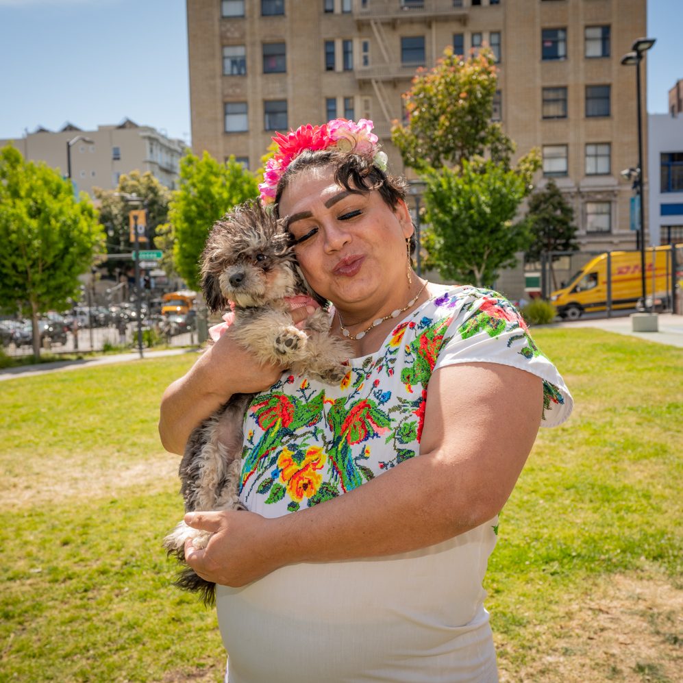 Johana (a woman) wearing a white dress with embroidered flowers and a flower headband, holding her small dog, standing in a park with buildings in the background.