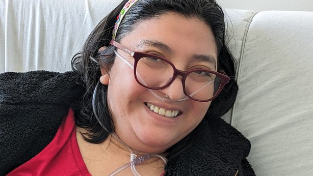 Edith, a woman wearing a red tee shirt, black hoodie, glasses, and a floral decorated headband, sitting up in her hospital bed with her oxygen tube, smiling.