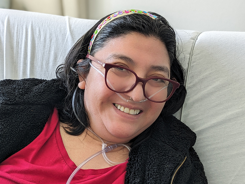 Edith, a woman wearing a red tee shirt, black hoodie, glasses, and a floral decorated headband, sitting up in her hospital bed with her oxygen tube, smiling.