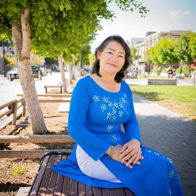 Cuc sitting on a park bench under trees, wearing a blue dress.